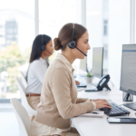 Une femme d'affaires portant un casque travaille à un bureau équipé d'un ordinateur, dans un espace de travail moderne et ouvert doté de grandes fenêtres. Elle tape sur le clavier tout en regardant l'écran.