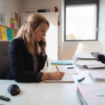 Une télésecrétaire blonde en veste noire à son bureau, en communication téléphonique tout en prenant des notes manuscrites sur un carnet
